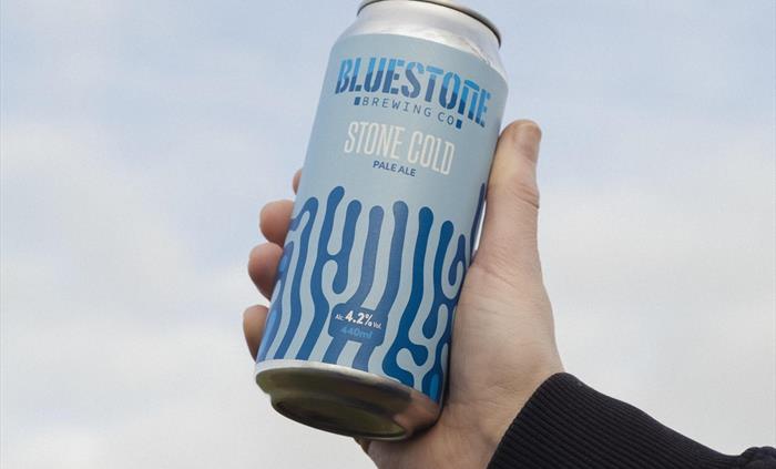 A hand holding up a can of beer to the sky, the label is blue with a swirly pattern on and read Stone Cold Pale Ale